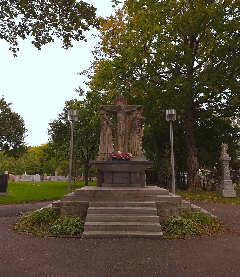 Stone calvary: Christ on the cross between two statues, on a stepped base, in the middle of a circle lined with tall trees.