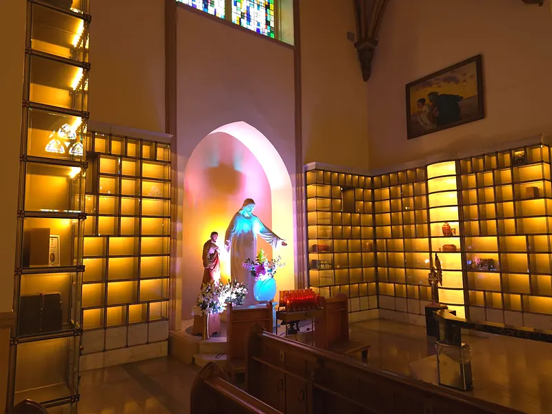 Glass niches bathed in golden light around a statue of Christ with open arms, beneath a stained-glass window, with pews in the foreground.