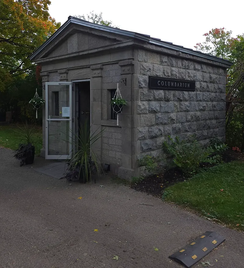 Small grey stone building with a pediment and a “Columbarium” plaque, its glass door open onto the lane.